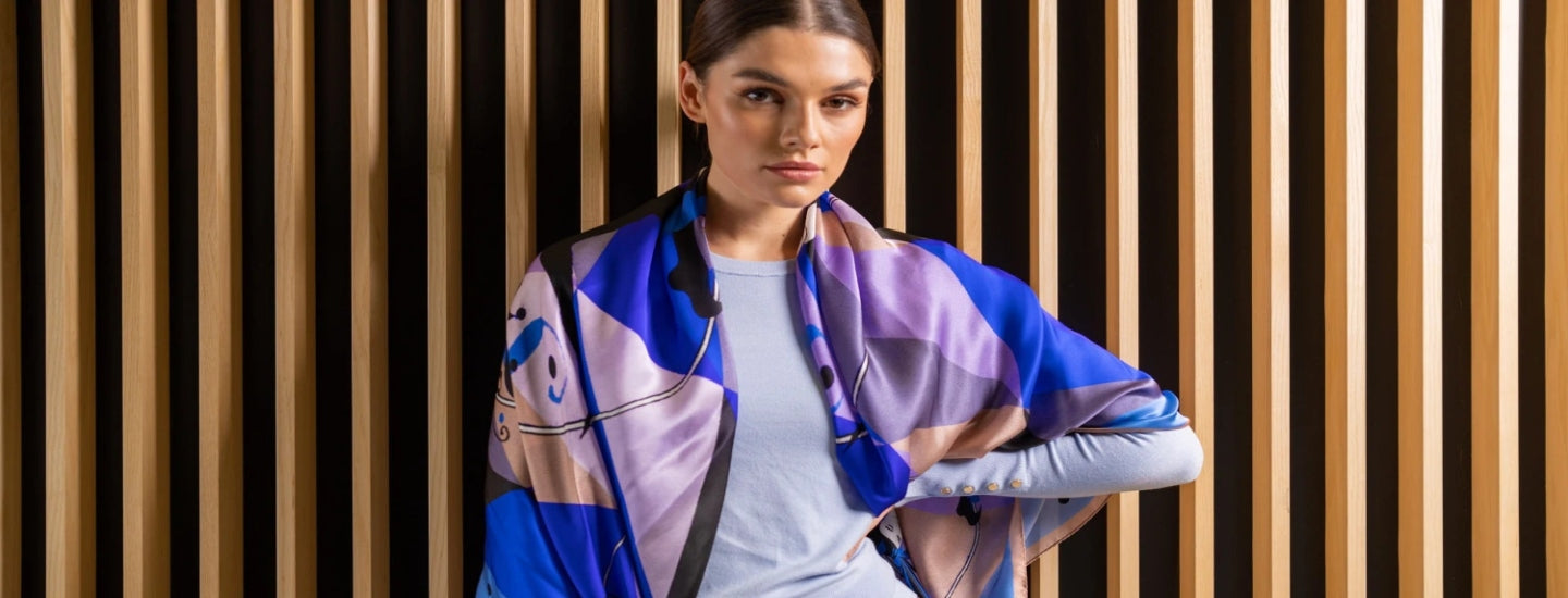 A woman stands in front of a slatted wooden wall, wearing a light blue shirt and a vibrant scarf with blue, purple, and beige geometric patterns draped over her shoulders. She looks directly at the camera with a confident expression.