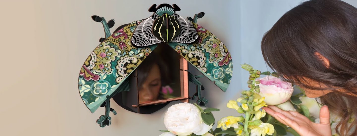 A person leans in with eyes closed to smell a large pink and white flower. Next to them on the wall is a decorative wall mirror with an ornate, colorful, beetle-shaped frame.