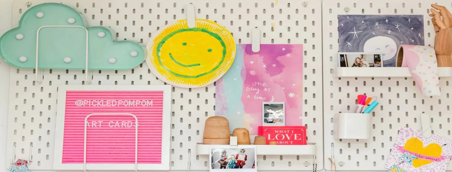 A white pegboard with various colorful items including a cloud light, a smiley face sun, a pink letter board with "@pickledpompon" and "ART CARDS" text, photos, a hand sculpture, markers, and decorative crafts.