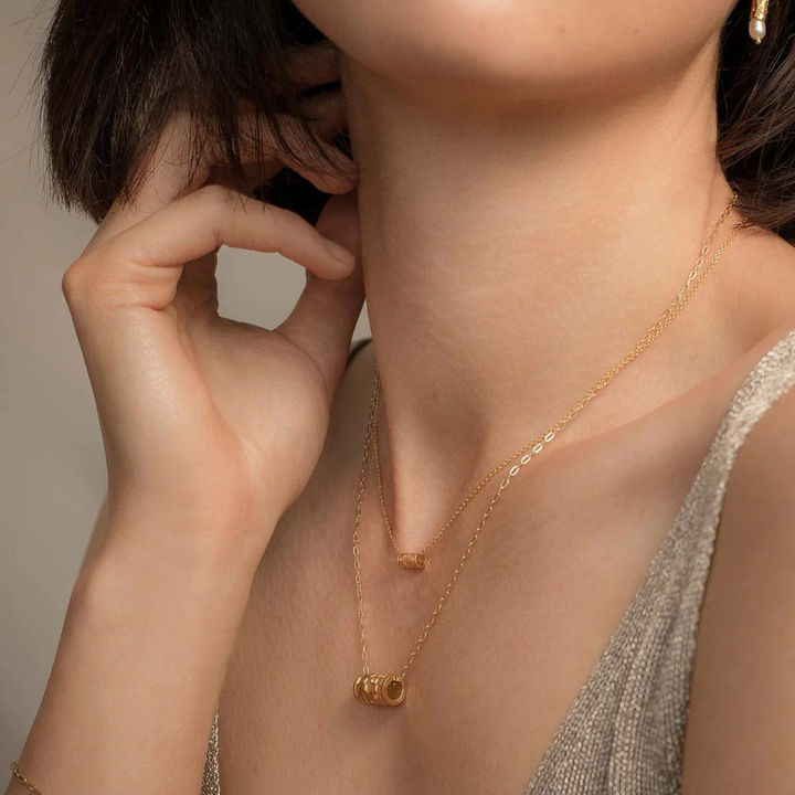 A short-haired individual wears Andrea Mears Jewellery's Tiny Karma Wheel Necklace Gold with a sleeveless shimmering top. One hand gently touches the necklace, creating an elegant and minimalistic look.