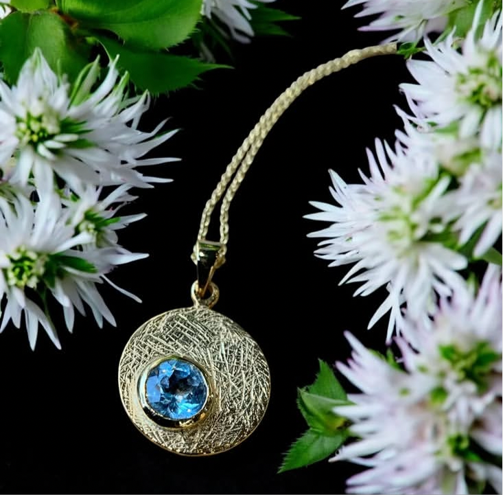 The Selene Necklace Gold - Small by Gallardo & Blaine Designs features a textured gold-plated circular pendant with a vibrant gemstone, beautifully showcased among delicate white and pale purple flowers on a black background.