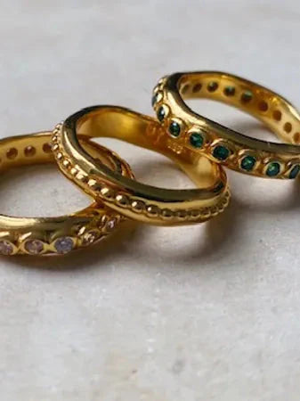 Three Shyla Astri Stacking Rings rest on a light surface: one gold ring with green stones, one with clear stones, and one featuring a beaded gold pattern.