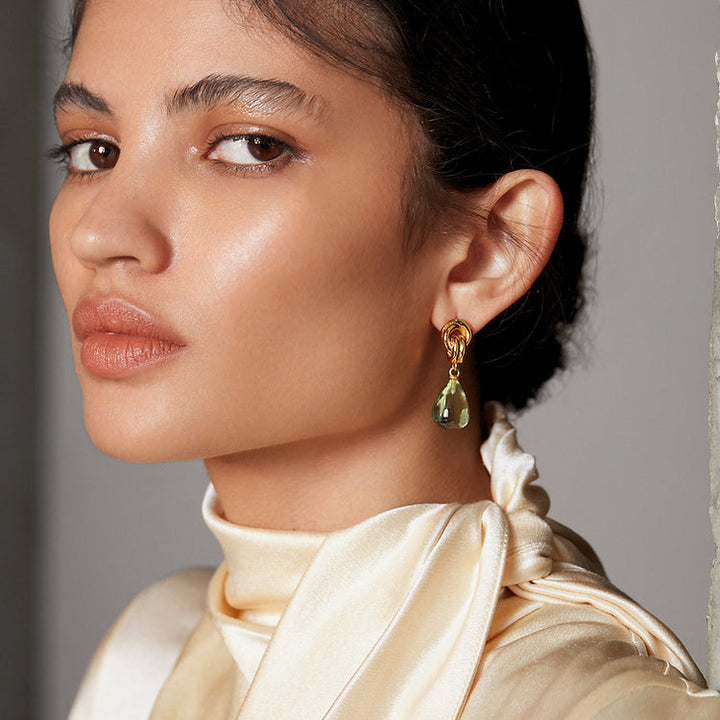 A person with smooth, glowing skin and neatly tied-back dark hair looks confidently into the distance. They are adorned with Shyla's Chunky Knot Earrings in high shine gold featuring a large green teardrop gem and are wearing a silky, cream-colored top with a stylish scarf-like collar.