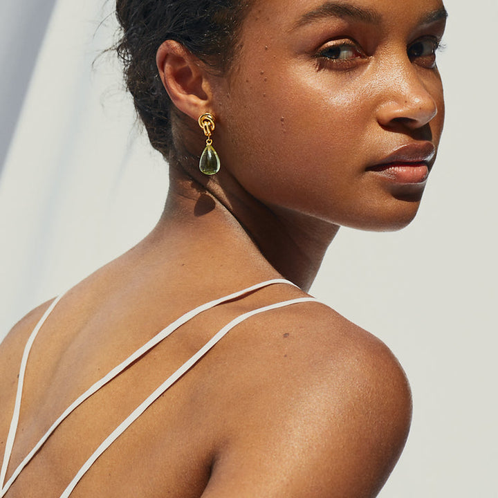 A woman with a bare back is turned slightly to the side, looking over her shoulder. She is wearing thin-strapped attire and Shyla’s high shine Chunky Knot Earrings adorned with green stones. The background is softly lit, highlighting her glowing skin and elegant pose.
