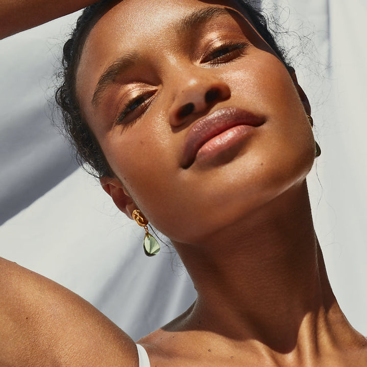 Close-up of a person with glowing skin, highlighted by natural sunlight. They are wearing Shyla's Chunky Knot Earrings in green and a white top. The background is blurred, showcasing a light fabric backdrop.