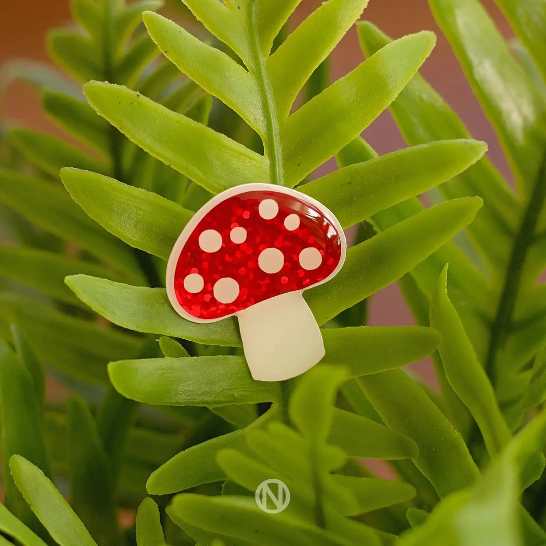 The Naoi Mushroom Pin Brooch features a red vinyl cap with white spots and a white stem, attached to green fern-like leaves, set against a blurred background.
