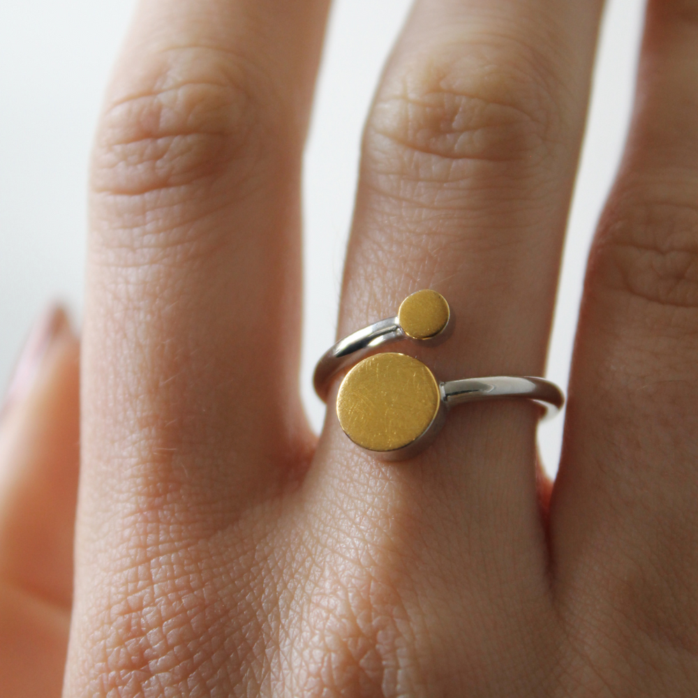 A hand displays the Gallardo & Blaine Designs Archeol Ring in sterling silver, featuring two round, flat gold vermeil accents of different sizes and an adjustable fit.