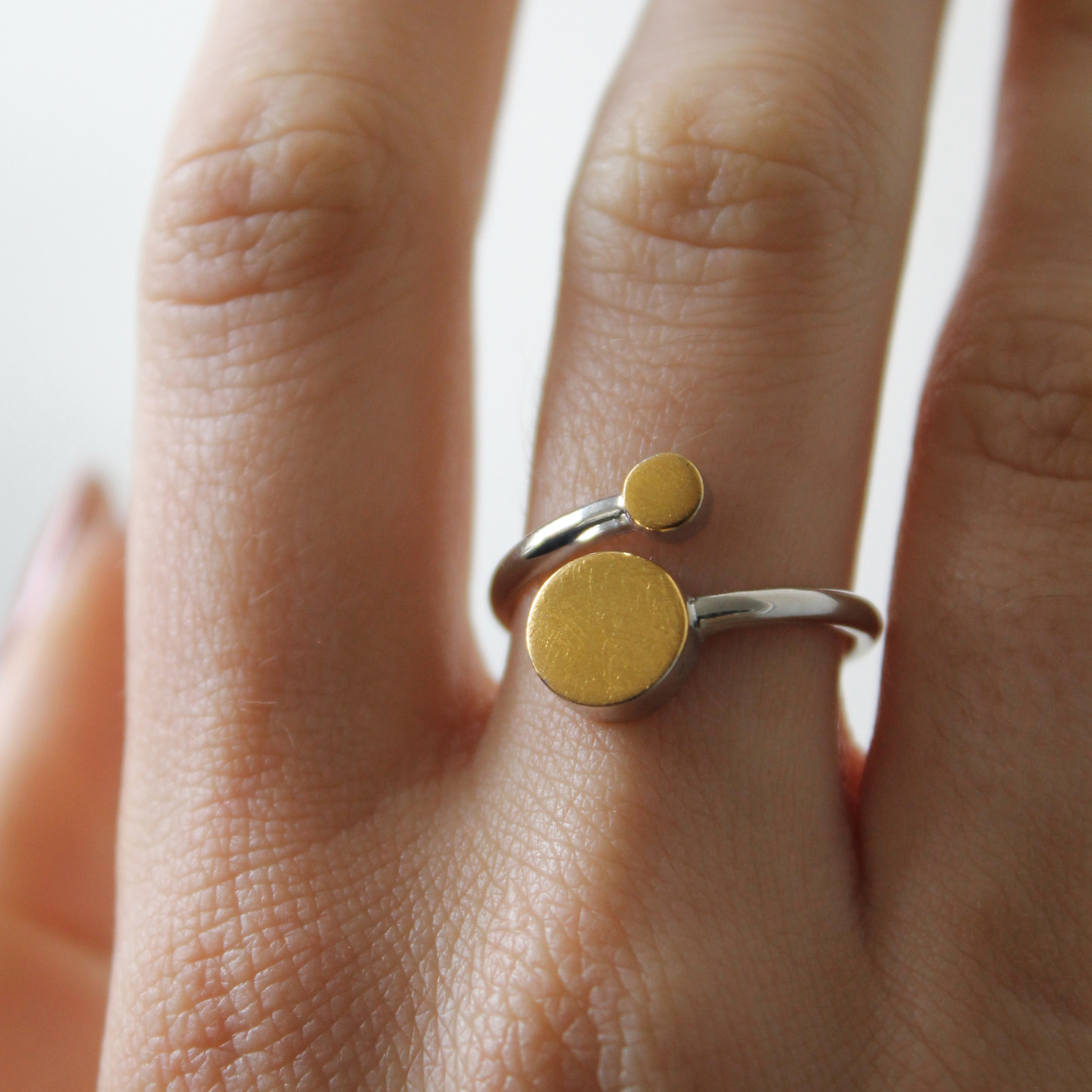 A hand displays the Gallardo & Blaine Designs Archeol Ring in sterling silver, featuring two round, flat gold vermeil accents of different sizes and an adjustable fit.