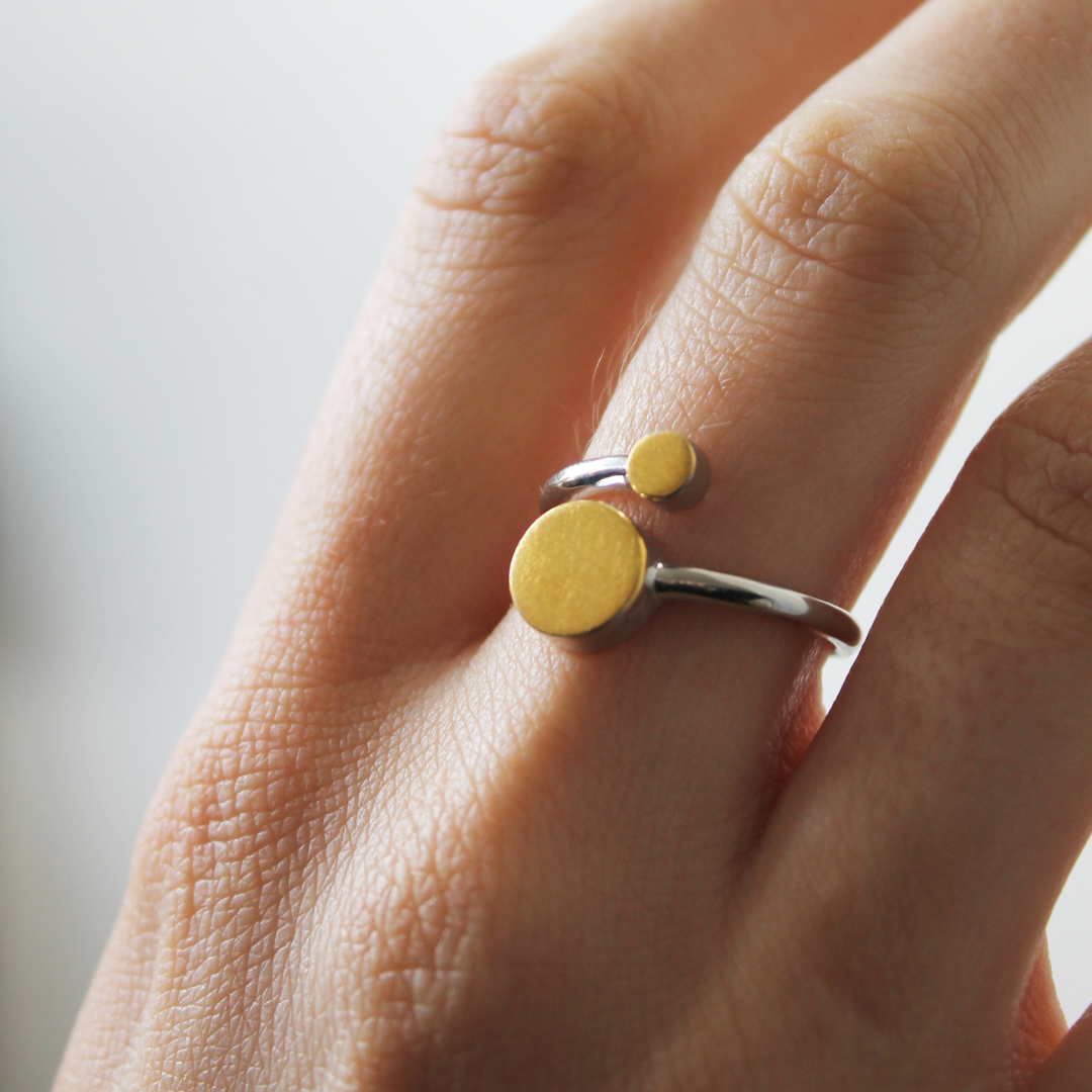 A close-up of a hand wearing the Archeol Ring by Gallardo & Blaine Designs, featuring adjustable sterling silver and two round, flat gold vermeil accents in different sizes. The softly blurred background enhances the elegant design.