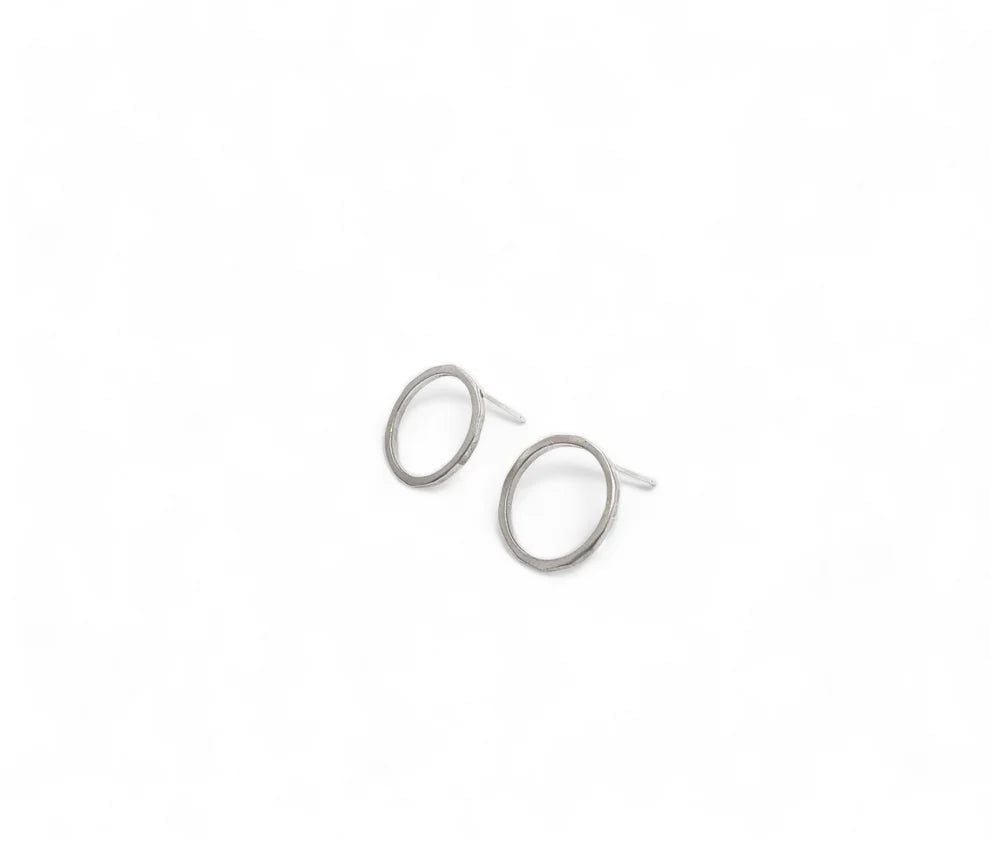 A pair of minimalist, hand-forged sterling silver hoop earrings against a plain white background. The small, elegant Ancaire Silver Studs by Lynsey de Burca have a simple circular design with thin bands and circle studs for securing them in place.