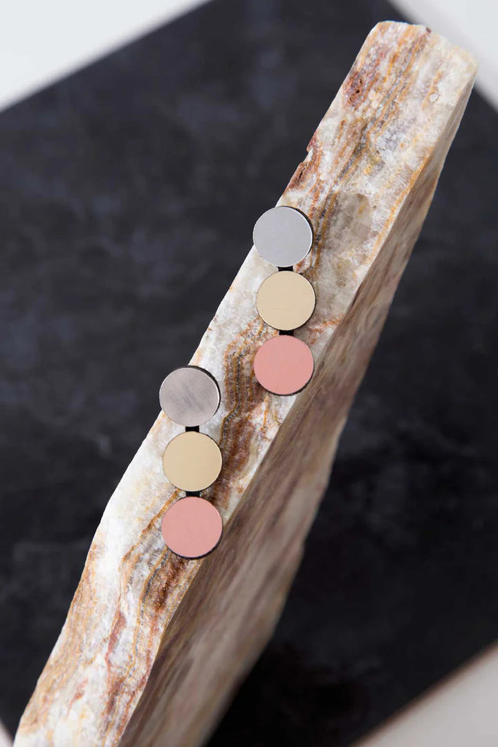 A pair of lightweight Abstraction 3 Dots Earrings by Iskin Sisters is displayed on the edge of a marble slab. The earrings feature four circular segments in varying colors: silver, gold, beige, and pink. The background is a contrasting dark surface, highlighting both the earrings and the slab.