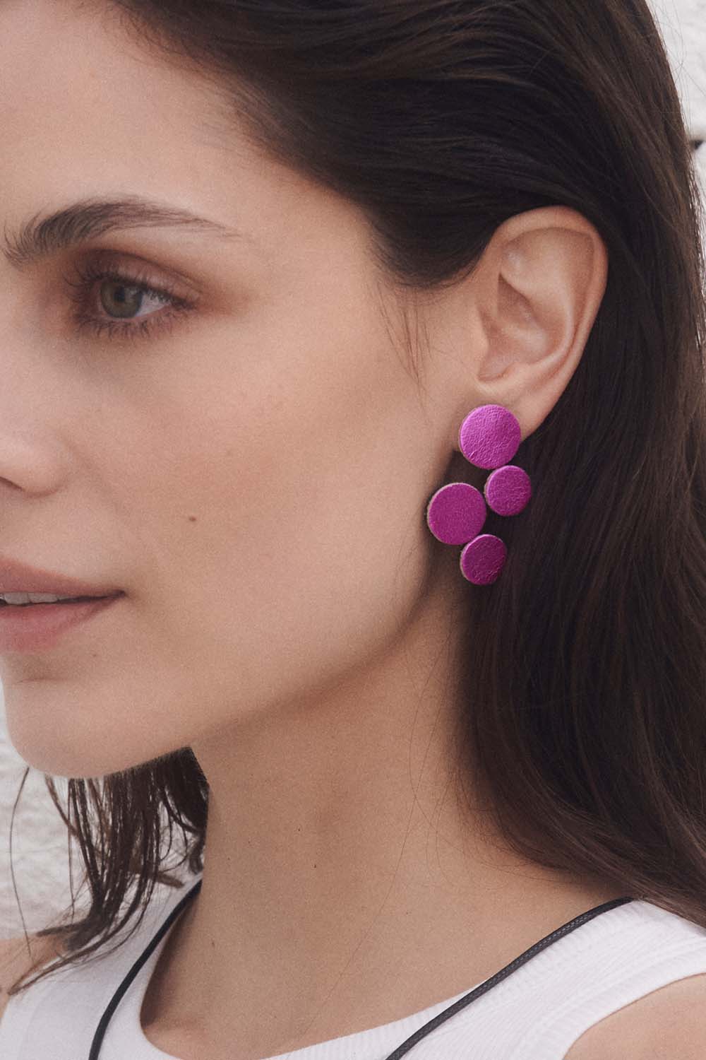 A woman with dark hair wears a white top and Iskin Sisters' Abstraction Pink Earrings, bold metallic fuchsia statement pieces that stand out against a neutral background as her makeup is highlighted in profile.