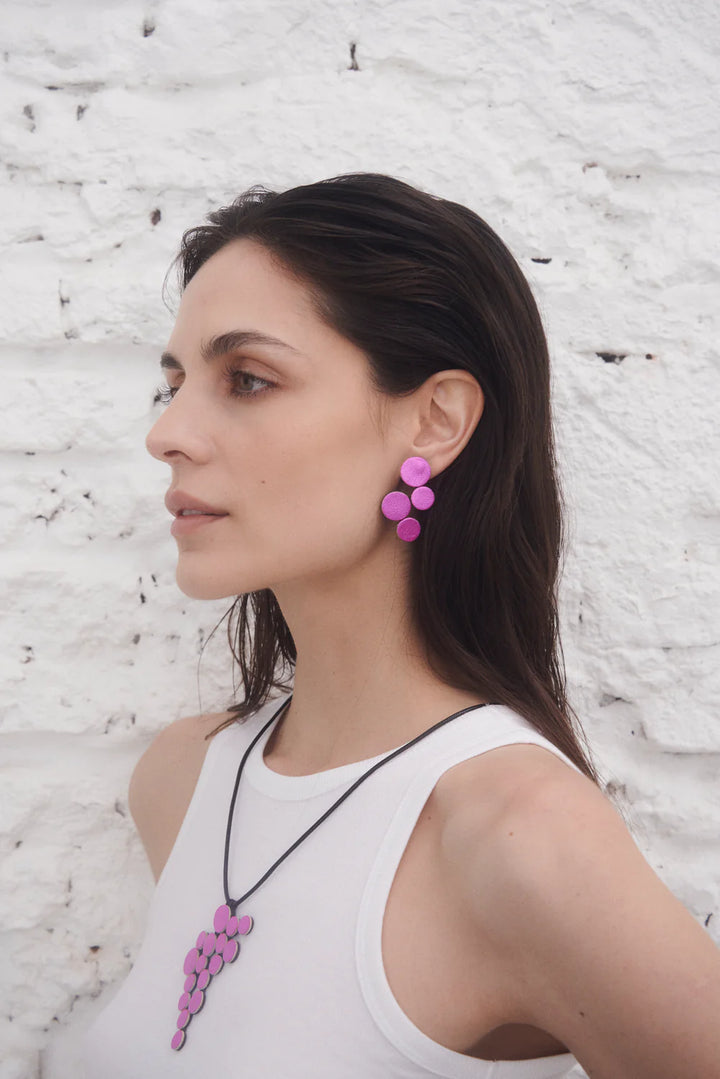 A woman with long dark hair wears a white sleeveless top and Iskin Sisters' Abstraction Pink Earrings. She stands in profile against a white textured brick wall.
