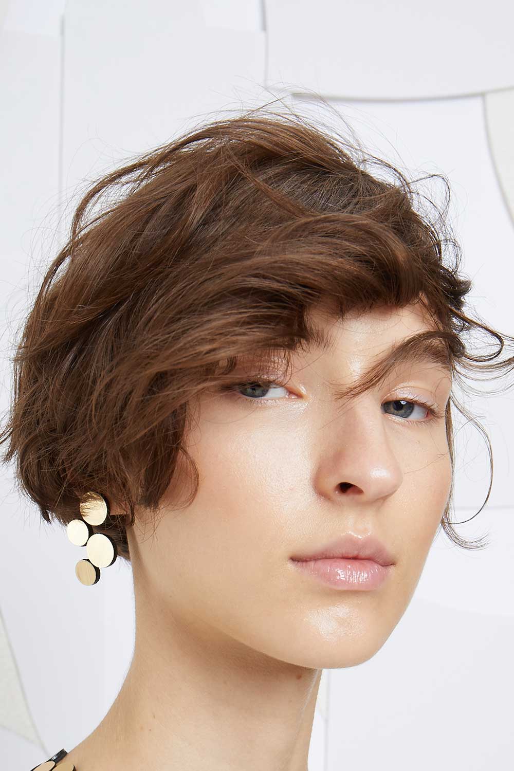A close-up of a person with short brown wavy hair and fair skin, wearing the Abstraction Gold Earrings by Iskin Sisters. They have a neutral expression, and the background is white with geometric shapes.