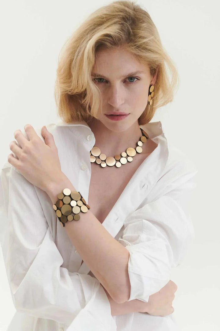 A blonde woman in a white shirt models Iskin Sisters' Abstraction Gold Earrings, paired with matching gold jewelry, against a light background.