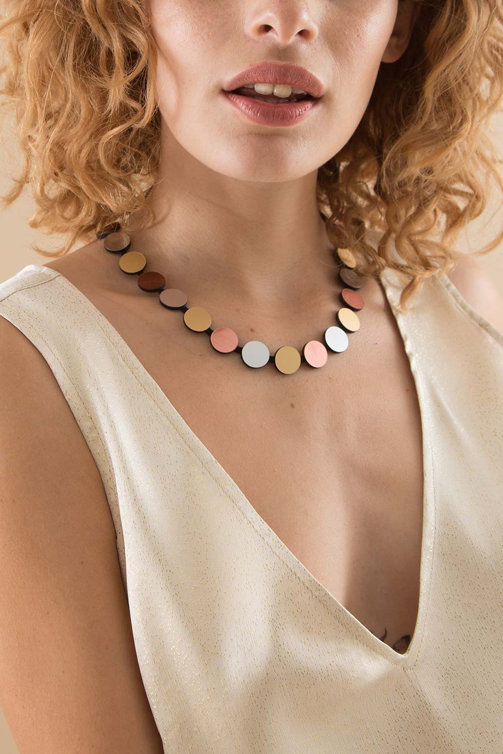 A woman with curly hair wears a cream deep V-neck top and the Abstraction Silver, Gold & Rose Gold Necklace by Iskin Sisters, featuring flat multicolored metal discs. The image is cropped to show her partial face and upper torso.