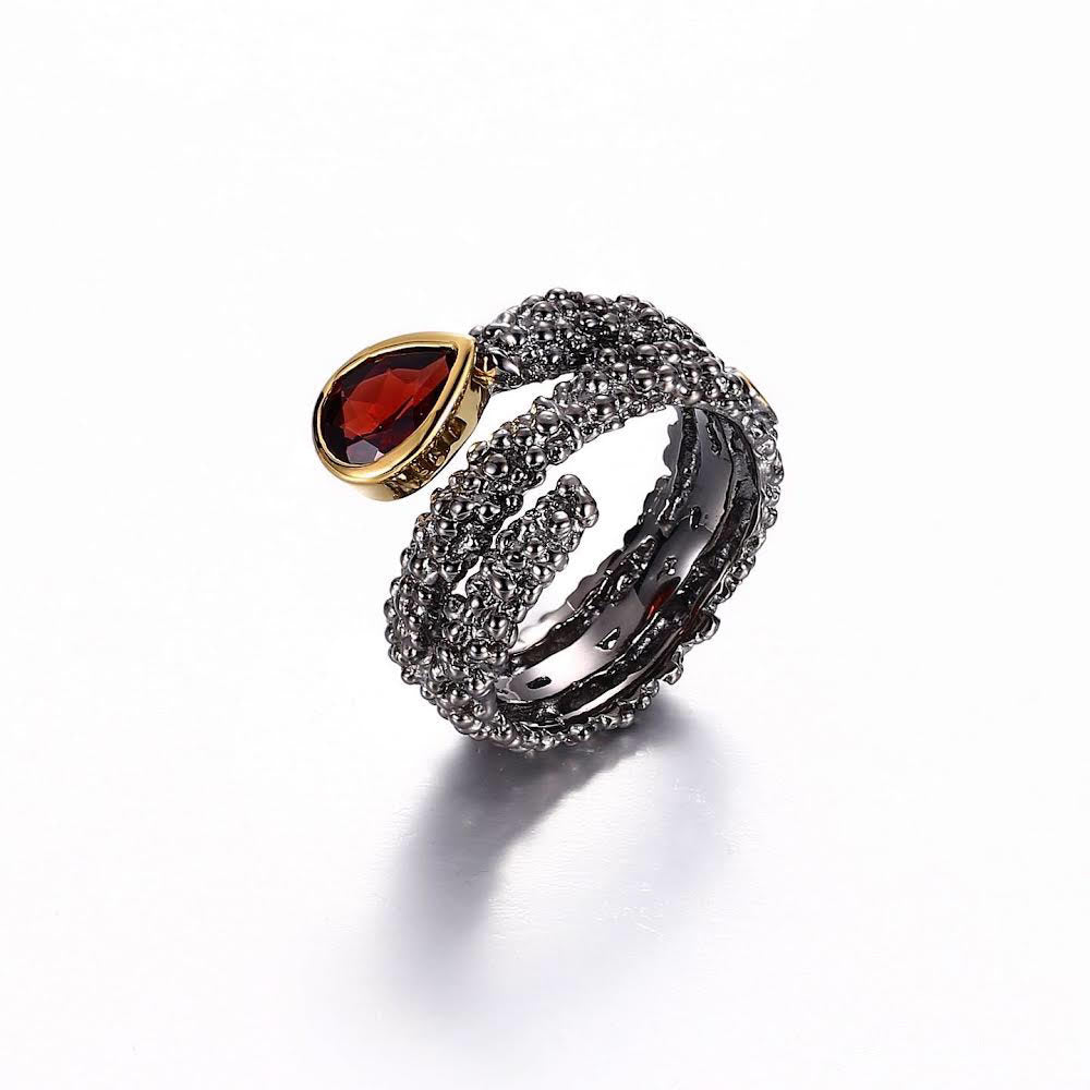 The Adelia Garnet Ring by Edith London features a coiled design of textured sterling silver, adorned with a pear-shaped, faceted red garnet stone set in gold. The intricately patterned surface shimmers brilliantly, displayed on a reflective white background.