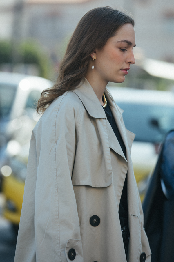 A woman with wavy brown hair wears a beige trench coat and Bonvo Agne Pearl Earrings while walking outdoors. The background is blurred, showing parked cars and greenery.
