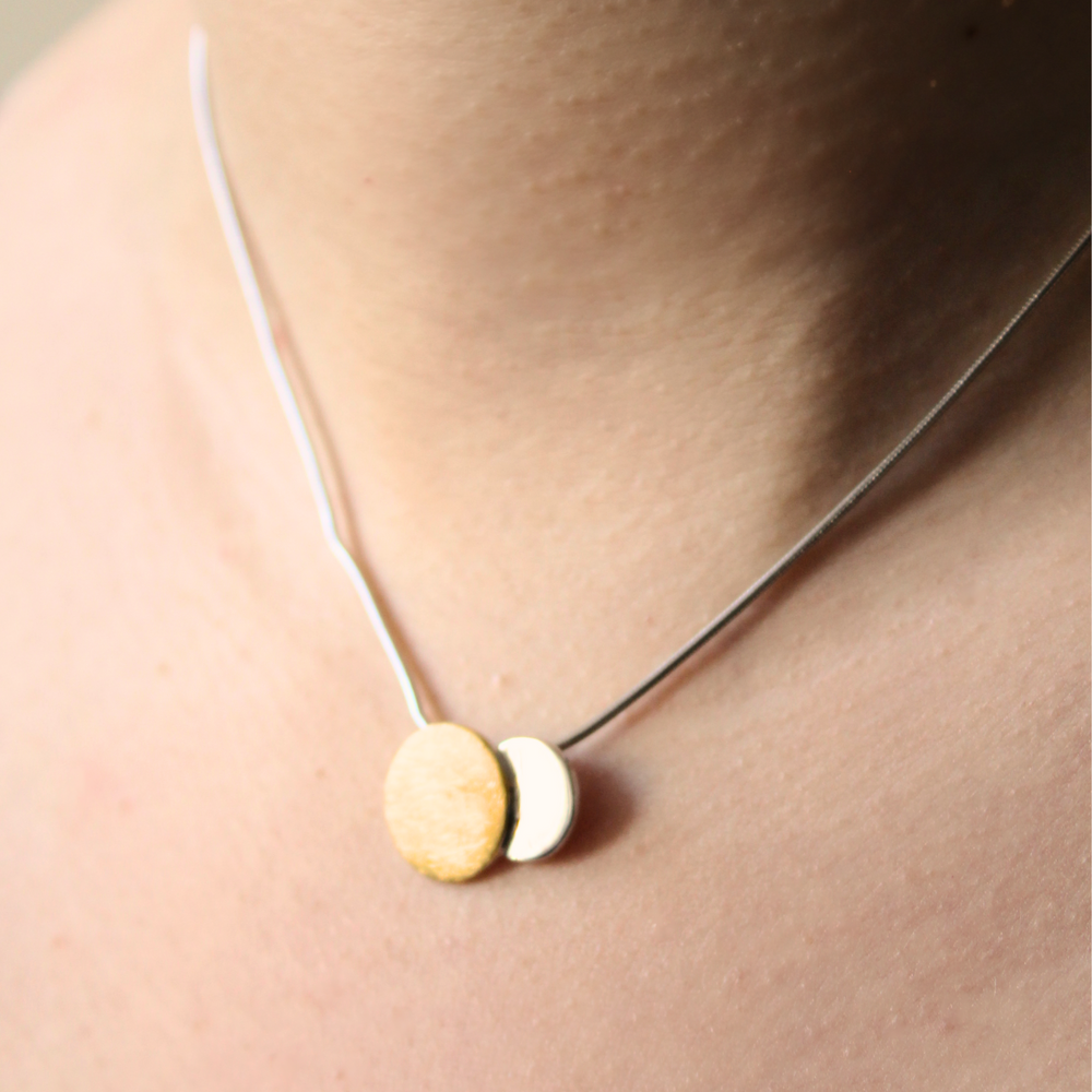 A close-up of a person's neck wearing the Gallardo & Blaine Designs Archeol Necklace in Sterling Silver & Gold Vermeil, featuring two round pendants—one gold vermeil and one white—side by side.