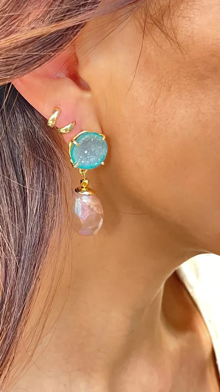 Close-up of a woman's ear wearing Aria Earrings by Aria V—featuring turquoise stones set in gold, a dangling translucent pink gemstone, and two small gold-plated brass ear cuffs—an elegant piece of handmade statement jewelry.