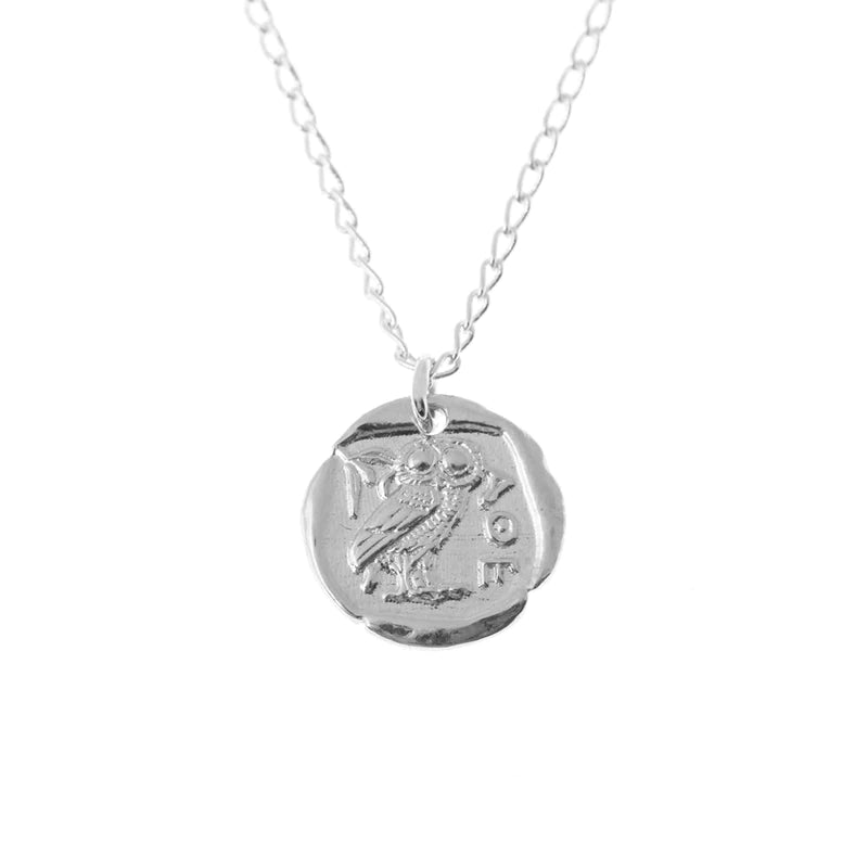 A sterling silver jewelry piece, the Andrea Mears Jewellery Athena Coin Necklace Silver features a round pendant with the embossed design of an owl and Greek letters. The pendant has a slightly irregular, ancient coin-like appearance, reminiscent of an Athena goddess pendant, and is connected to a delicate silver chain.
