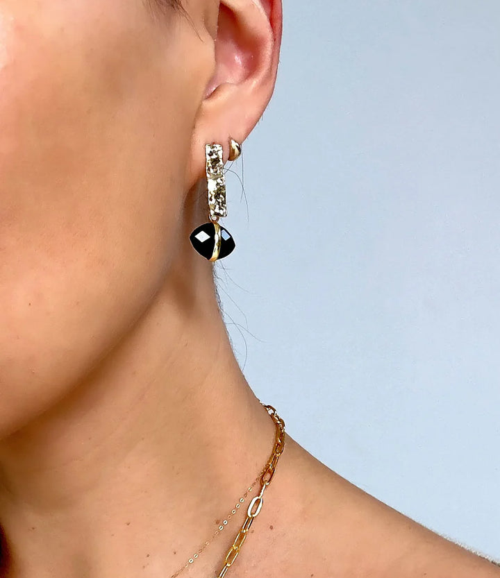 Close-up of a woman wearing Aria V's Ayin Earrings—handcrafted, gold plated with clear gemstones and a black geometric pendant—paired with layered gold necklaces against a light blue background.