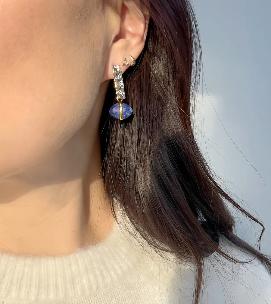 A close-up of a person with long brown hair, wearing a light sweater and Ayin Earrings by Aria V—handcrafted silver hoops with gold-plated accents and a blue natural stone.
