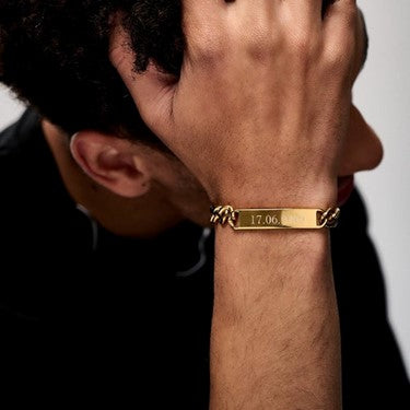 A person touches their forehead with their hand, displaying a Chunky Chain ID Bar Bracelet - Orelia & Joe on their wrist. The gold bracelet features an engraved date "17.06." The person has short, curly hair and is wearing a dark shirt. The background is a plain, light color.