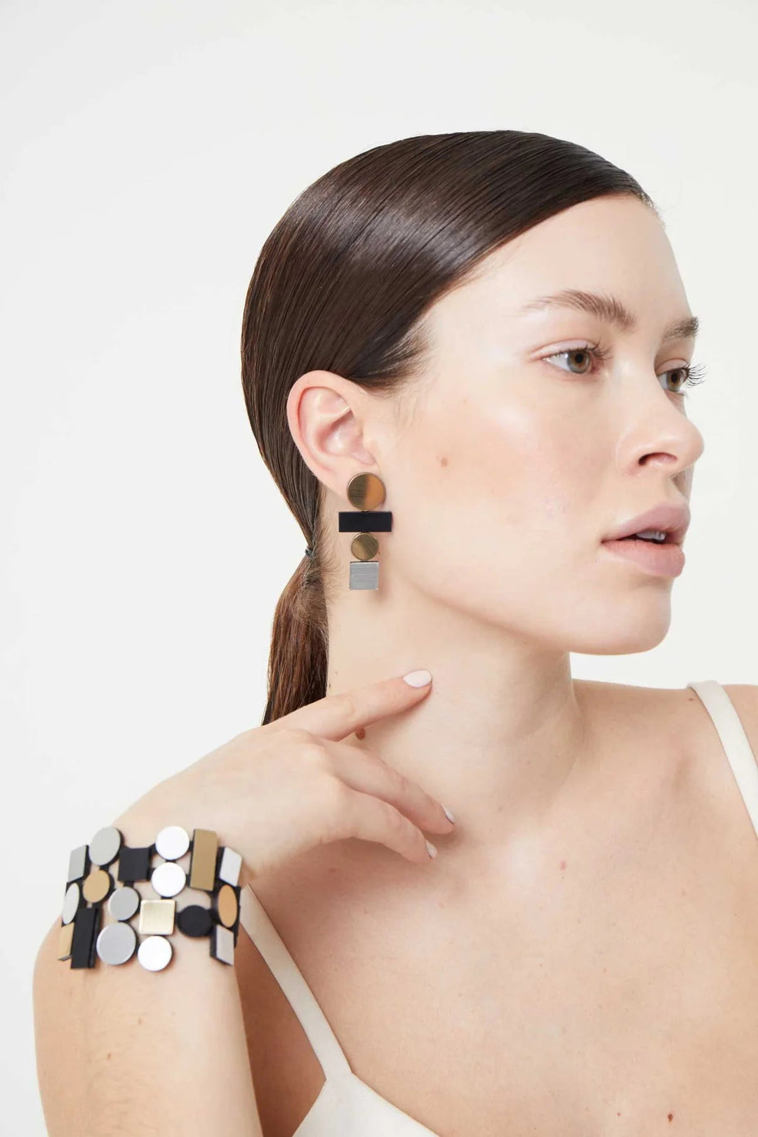 A woman with long, straight hair pulled back wears large geometric-patterned earrings from the Bauhaus Collection by Iskin Sisters and a matching bracelet. She is dressed in a white sleeveless top and poses with her left hand near her face, looking to the side against a plain, light-colored background.
