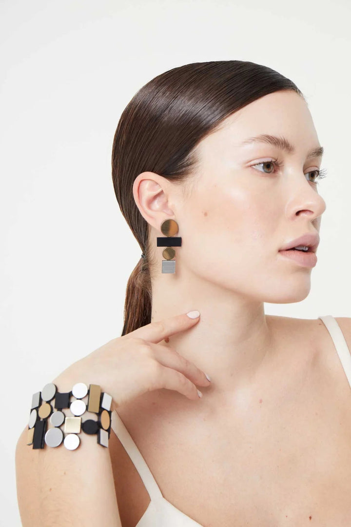 A woman with long, straight hair pulled back wears large geometric-patterned earrings from the Bauhaus Collection by Iskin Sisters and a matching bracelet. She is dressed in a white sleeveless top and poses with her left hand near her face, looking to the side against a plain, light-colored background.