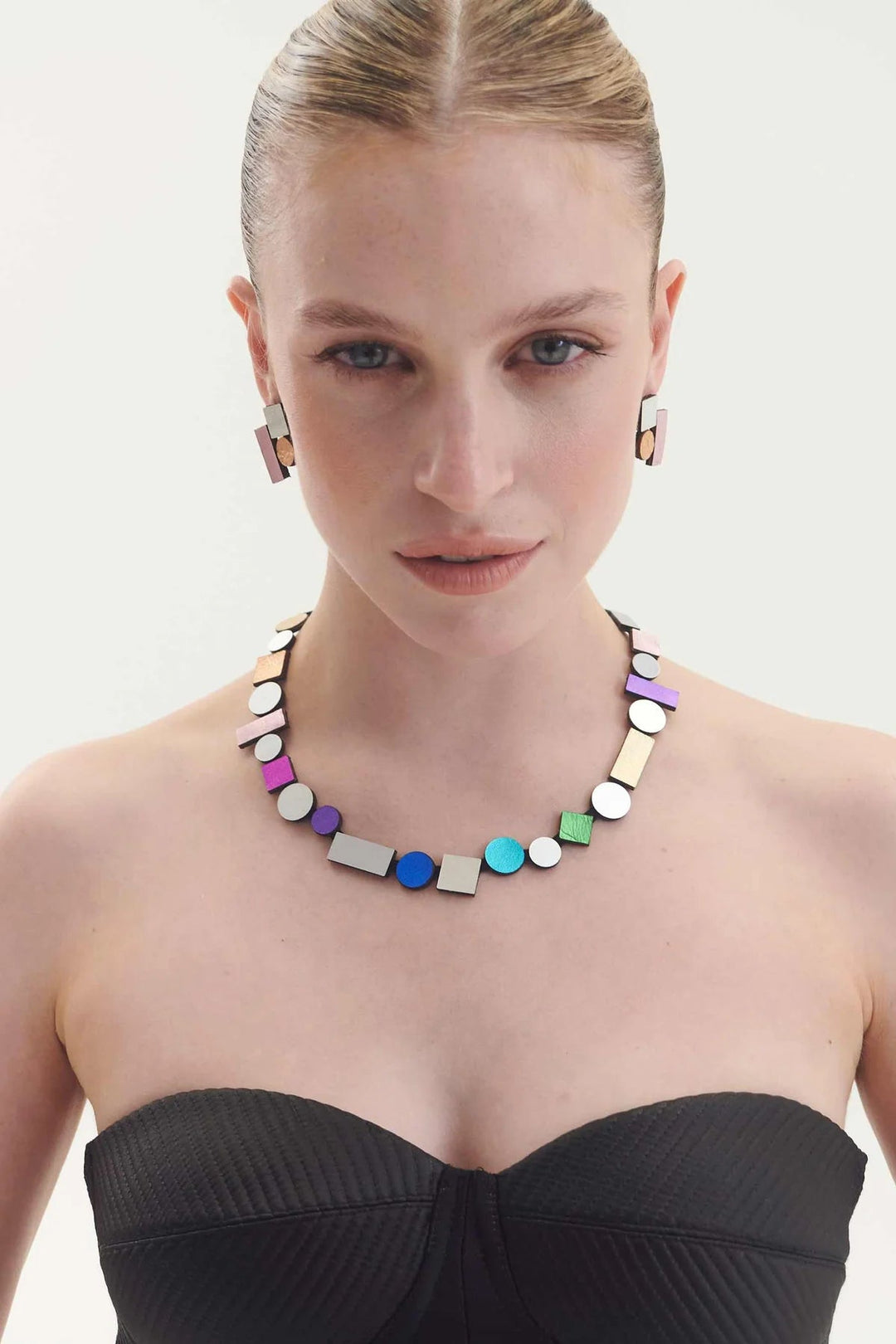 A woman with blonde hair styled in a sleek bun is wearing a strapless black top. She accessorizes with the Bauhaus Rainbow Necklace and matching earrings from Iskin Sisters' collection, featuring colorful shapes in blue, purple, green, and metallic tones. She poses against a plain white background.
