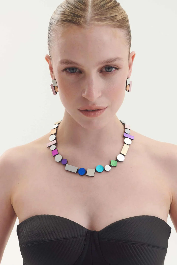 A woman with slicked-back hair wears geometric earrings and the Iskin Sisters' Bauhaus Rainbow Necklace, featuring colorful shapes, paired with a black strapless top against a plain background.