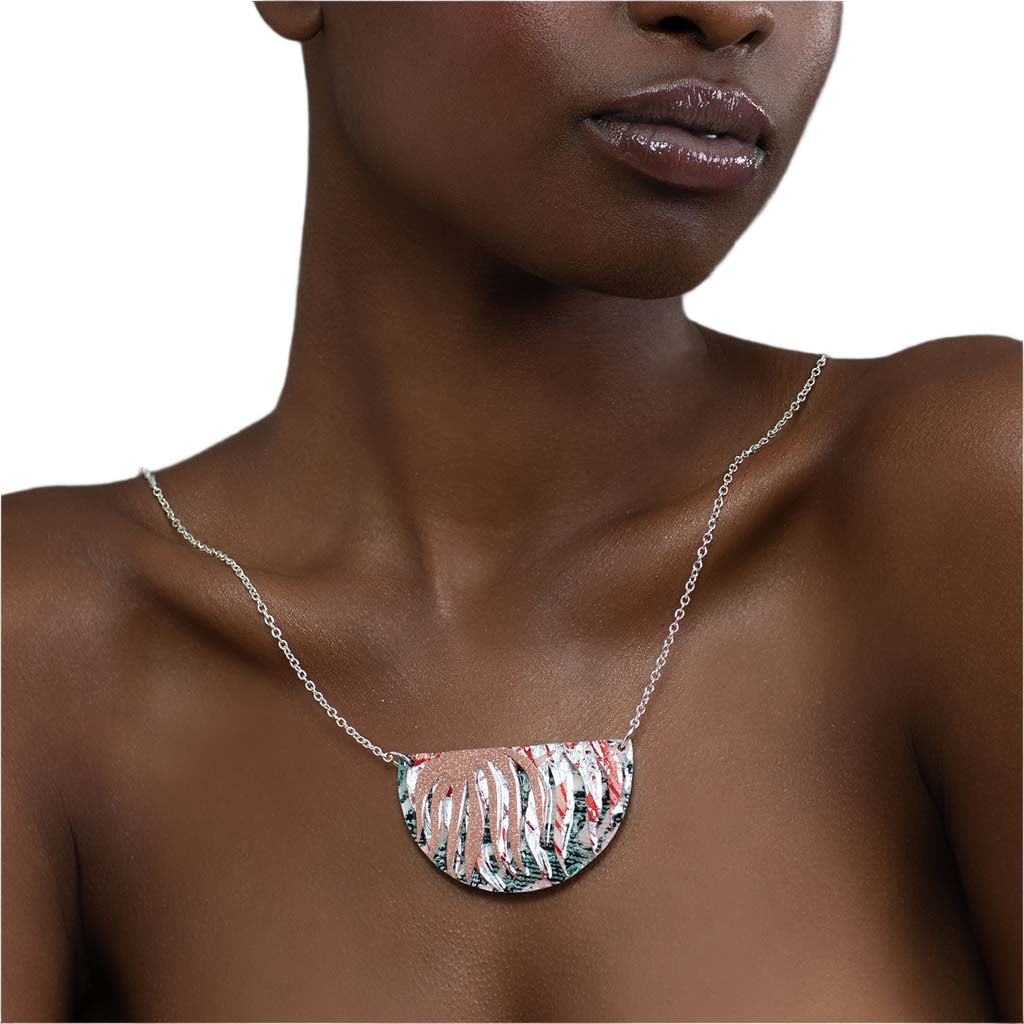 A woman with bare shoulders wears the Rothlú Bláth Batik Textile Necklace in Dark-Green/Blush Shimmer/Silver/Coral, featuring a large semi-circular pendant with abstract cut-outs. The white background highlights this unique handmade jewelry piece.