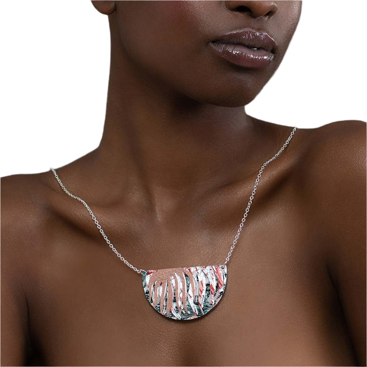 A woman with bare shoulders wears the Rothlú Bláth Batik Textile Necklace in Dark-Green/Blush Shimmer/Silver/Coral, featuring a large semi-circular pendant with abstract cut-outs. The white background highlights this unique handmade jewelry piece.