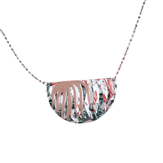 The Rothlú Bláth Batik Textile Necklace features a thin chain and a semicircular pendant with metallic leaf over dark-green, blush shimmer, silver, and coral—perfect for fans of distinctive handmade jewelry.