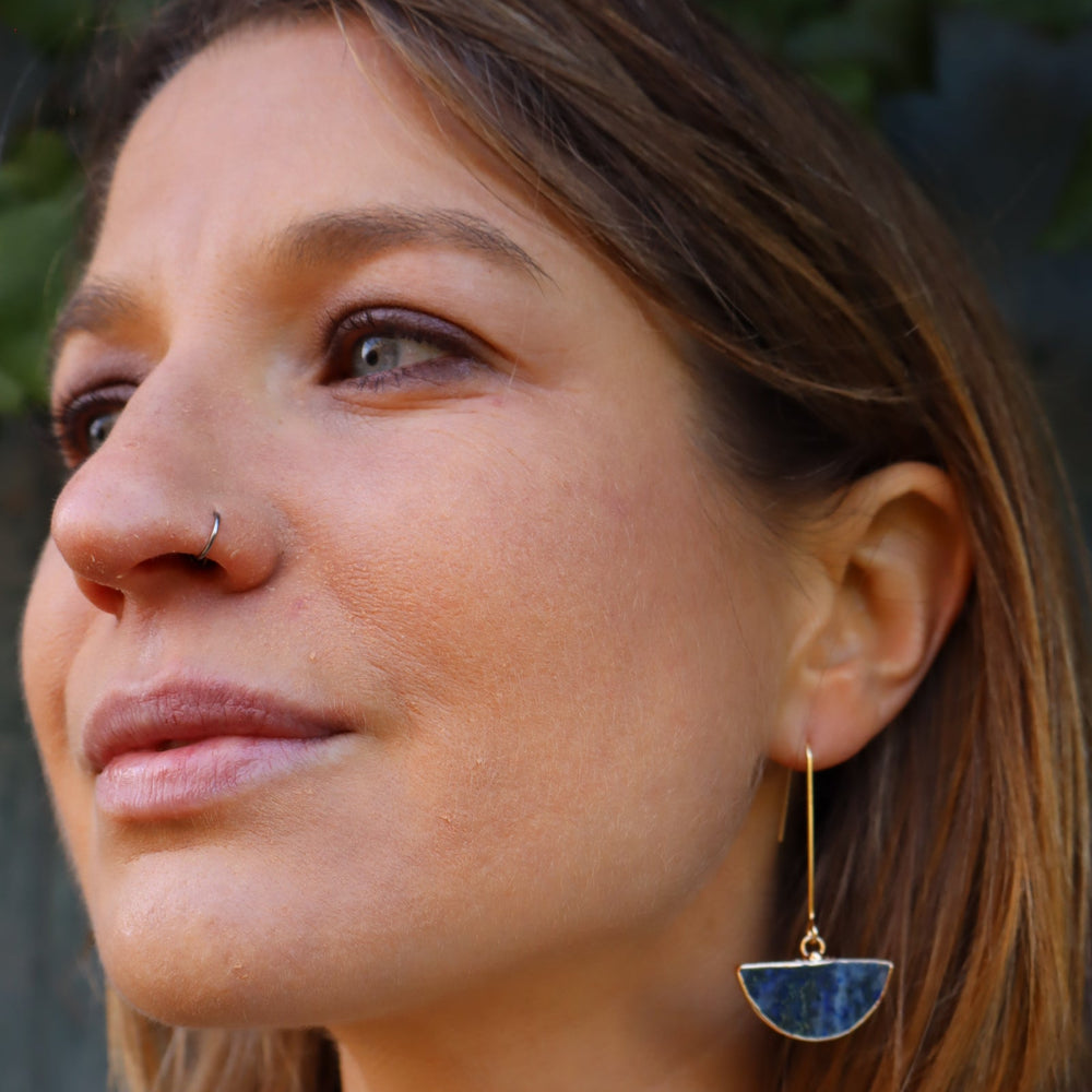 A person with long hair is shown in close-up, wearing a nose ring and Blue Moon Lapis Earrings from Kybalion Jewellery. The handmade, gold-plated earrings feature semicircles with blue and gold details. They gaze upward against a slightly blurred backdrop of greenery.