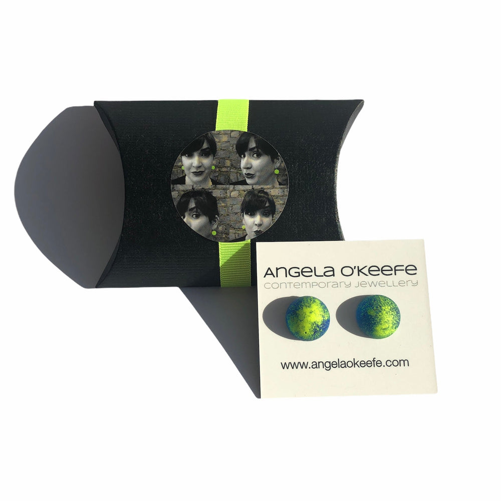 A black, cylindrical gift box with a neon green ribbon, featuring a circular image of a woman's face in four sections. Beside it is a business card with vibrant blue and green Graffiti Stud earrings and the text: "Angela O'Keefe Contemporary Jewellery" and a website link. The bright colors add an eye-catching touch.