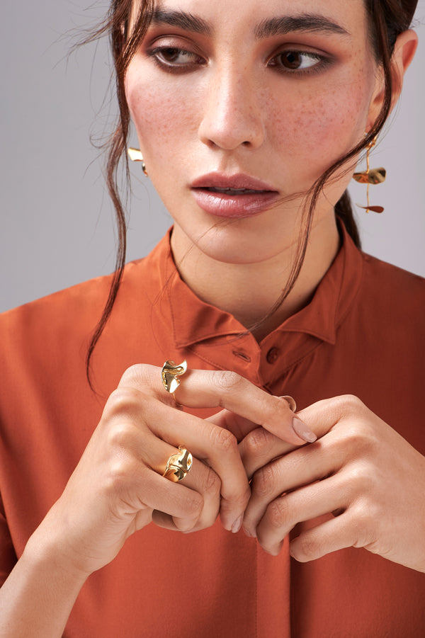 A woman in a rust blouse wears bold gold-plated earrings and two chunky Bonvo Flor Rings, looks slightly away with her hands gently touching. She has natural makeup and freckles.