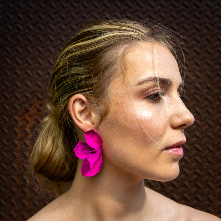 A woman with blonde hair tied back is shown in profile against a textured, dark background, wearing a striking handmade bright pink Bougainvillea Earring by Kybalion Jewellery. Her natural makeup complements her unique and nickel-free look.