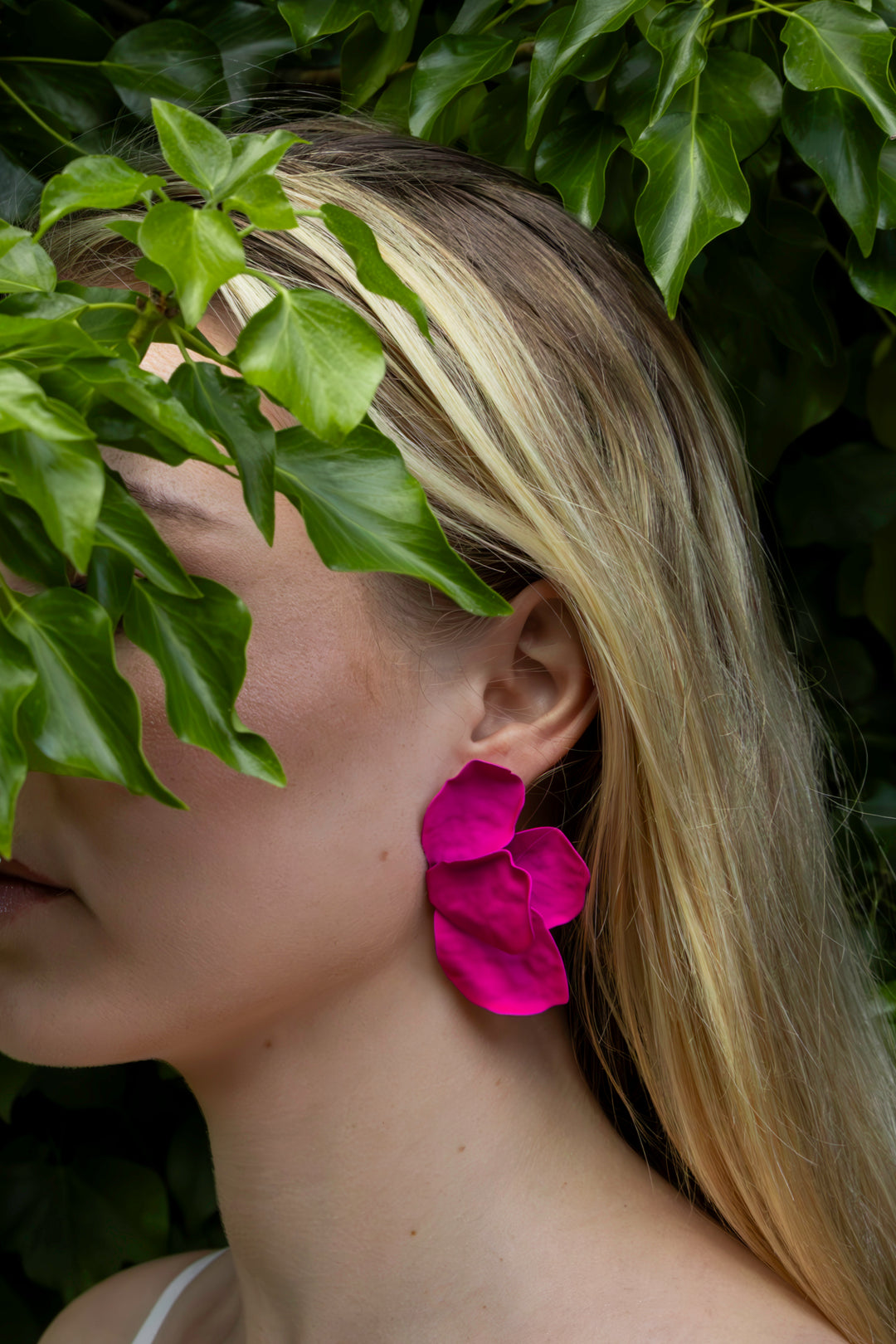 A woman with long blonde hair, partially hidden by lush green leaves, wears Kybalion Jewellery's Bougainvillea Earrings in bright pink. The earrings' handmade flower shape and nickel-free design create a vibrant contrast against the natural backdrop.