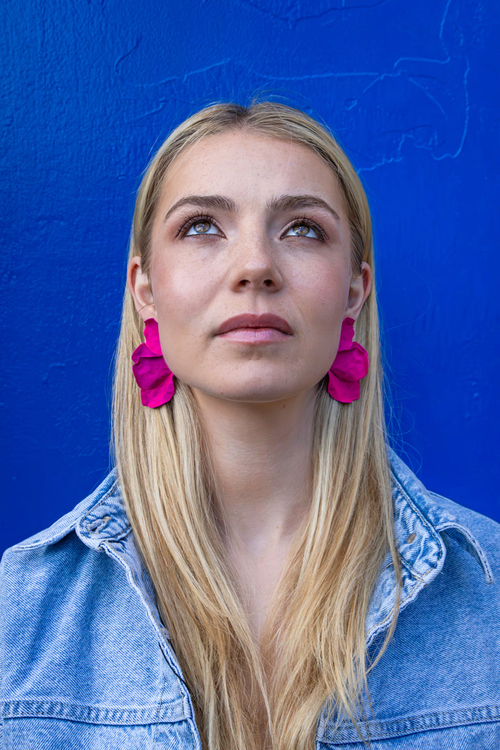 A person with long blonde hair gazes upwards, wearing Kybalion Jewellery's Bougainvillea Earrings in bright pink, which are handmade and nickel-free. They also wear a light denim jacket and stand against a vibrant blue background.