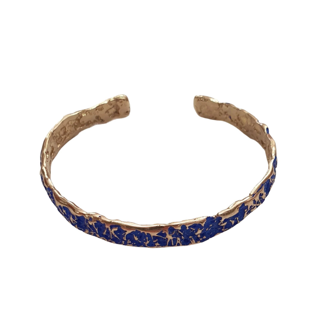 A Crackle Texture Coloured Bangle by Georgia Charal, with a crackle texture finish, featuring a central inlay of vibrant blue material, possibly enamel or stones, that has an irregular, organic pattern. Handmade in Athens, the open-ended bracelet rests elegantly on a white surface.