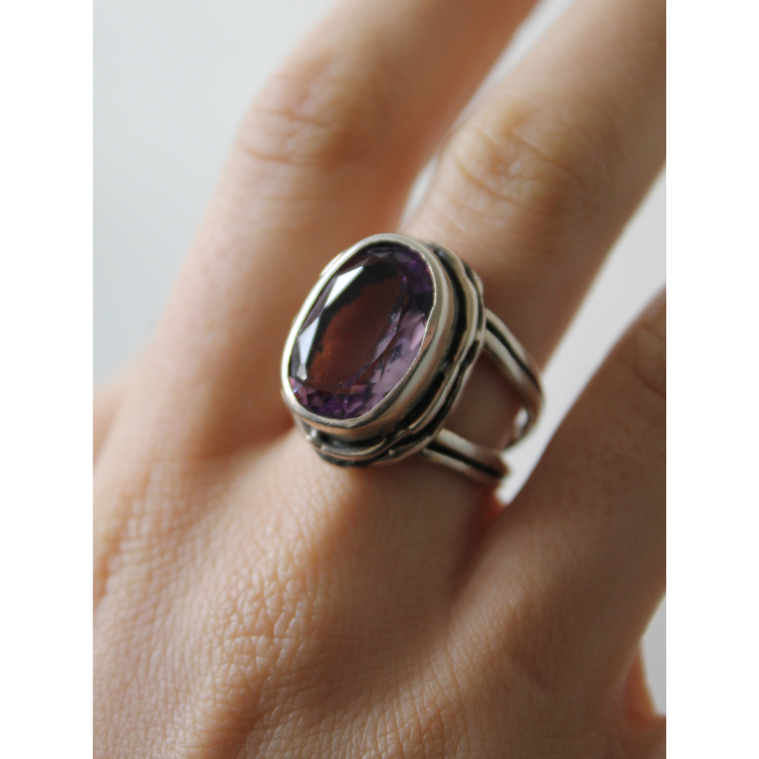 A hand wears the Magnolia Ring by Gallardo & Blaine Designs, featuring sterling silver, a large oval purple gemstone, a double band, and detailed metalwork for added elegance.