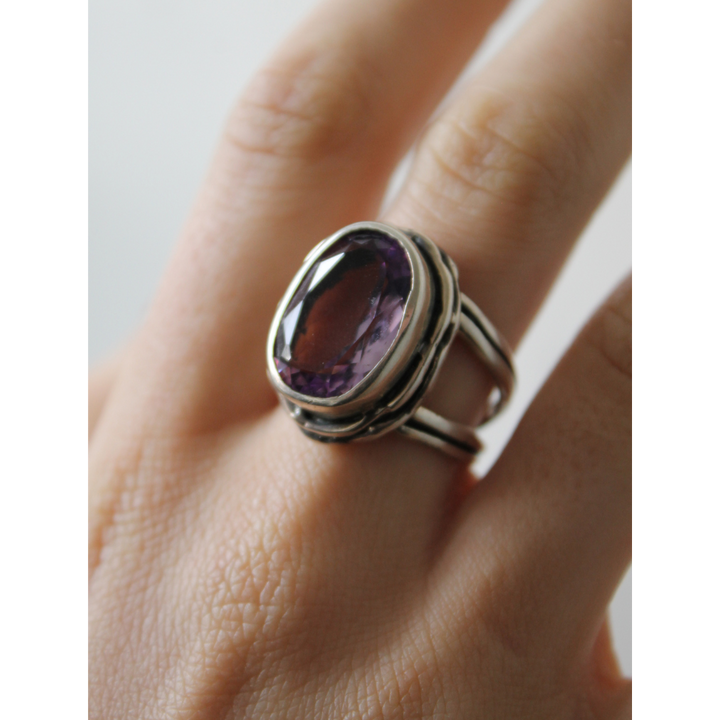 A hand wears the Magnolia Ring by Gallardo & Blaine Designs, featuring sterling silver, a large oval purple gemstone, a double band, and detailed metalwork for added elegance.