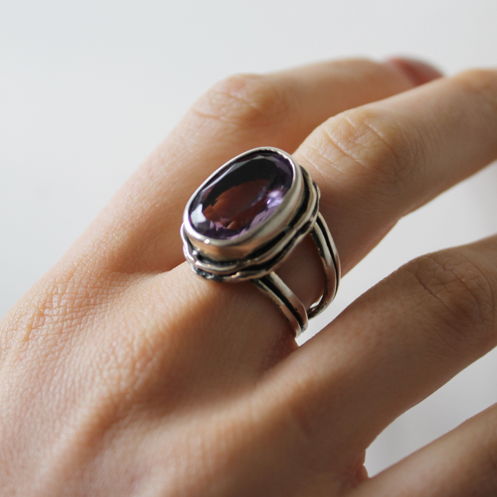 A hand wears the Magnolia Ring by Gallardo & Blaine Designs, crafted in silver-gold and set with a large purple gemstone, displayed against a softly blurred background.