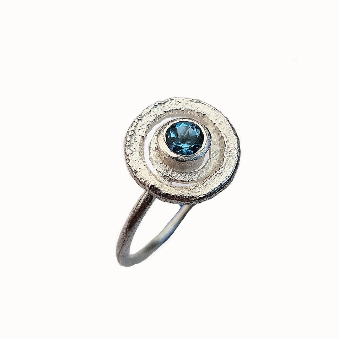 The Caroline Stokesberry-lee Blue Topaz Planetary Ring features a textured silver circular design with a blue topaz gemstone in the center, shown on a white background.
