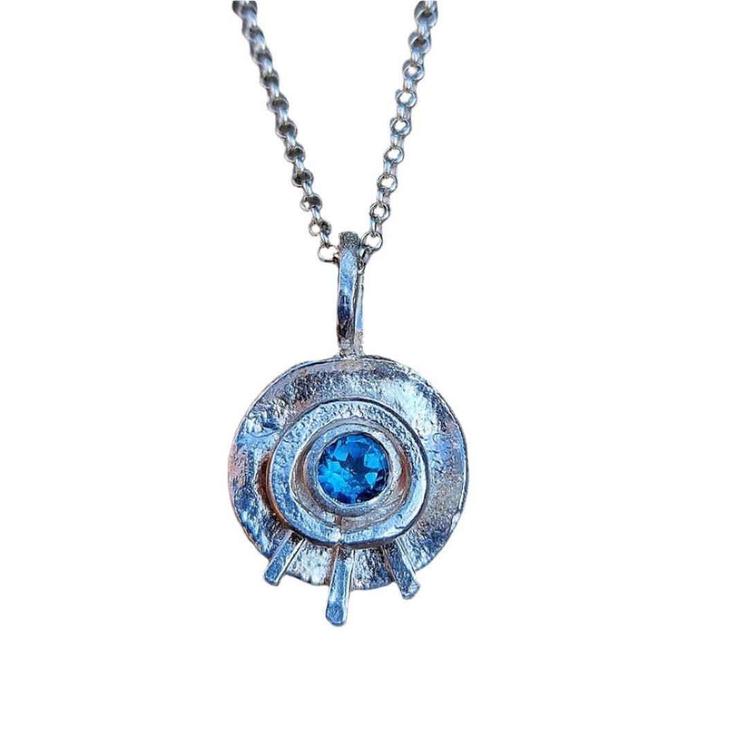 Introducing the Celestial Eye Pendant by Caroline Stokesberry-lee, a stunning sterling silver piece featuring a textured circular design with a vibrant blue topaz gemstone at its center, enhanced by elegant radial lines.