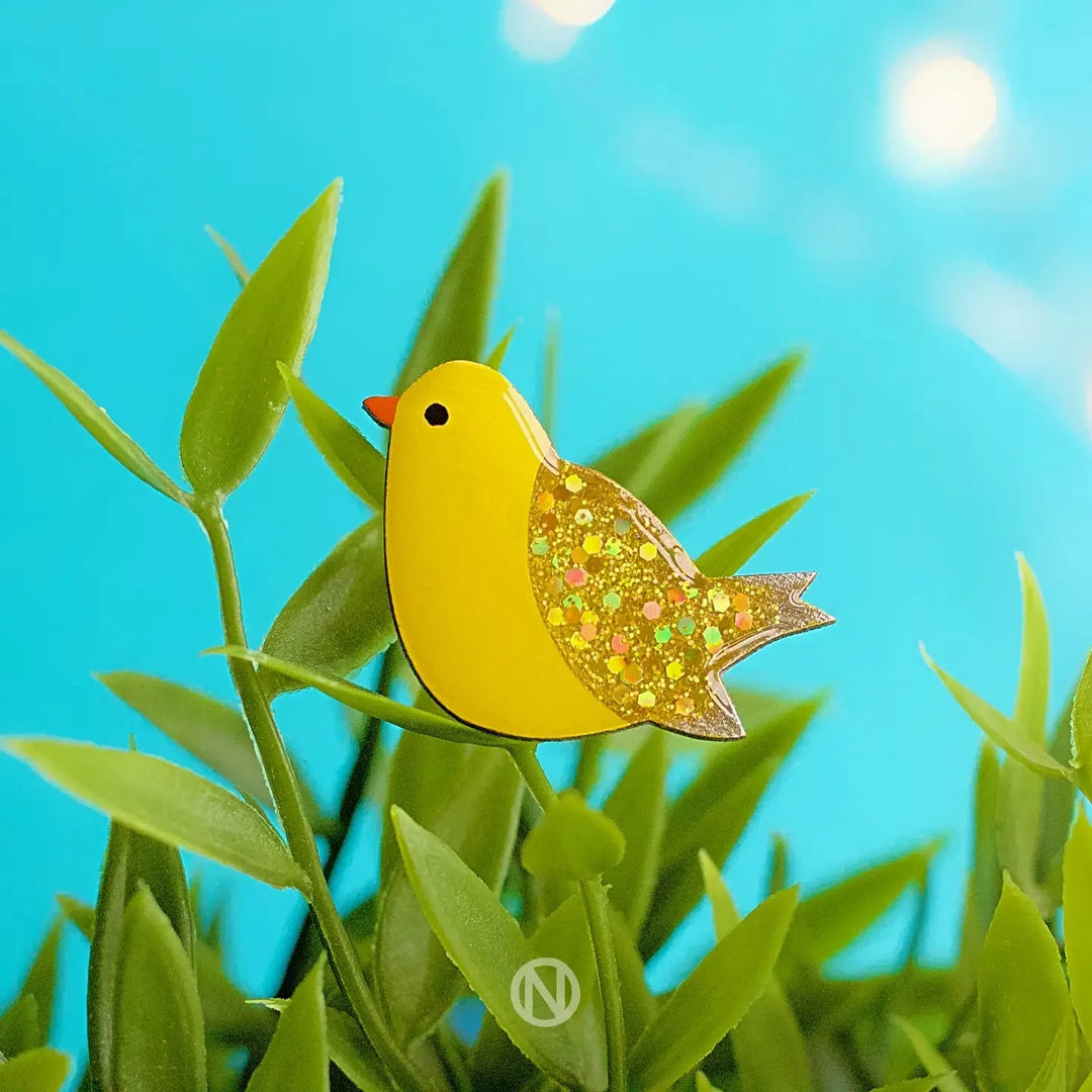 The Naoi Canary Pin Brooch is a hand-painted wooden bird with a glittery wing, displayed on green leaves against a bright blue sky and soft sunlight in the background.