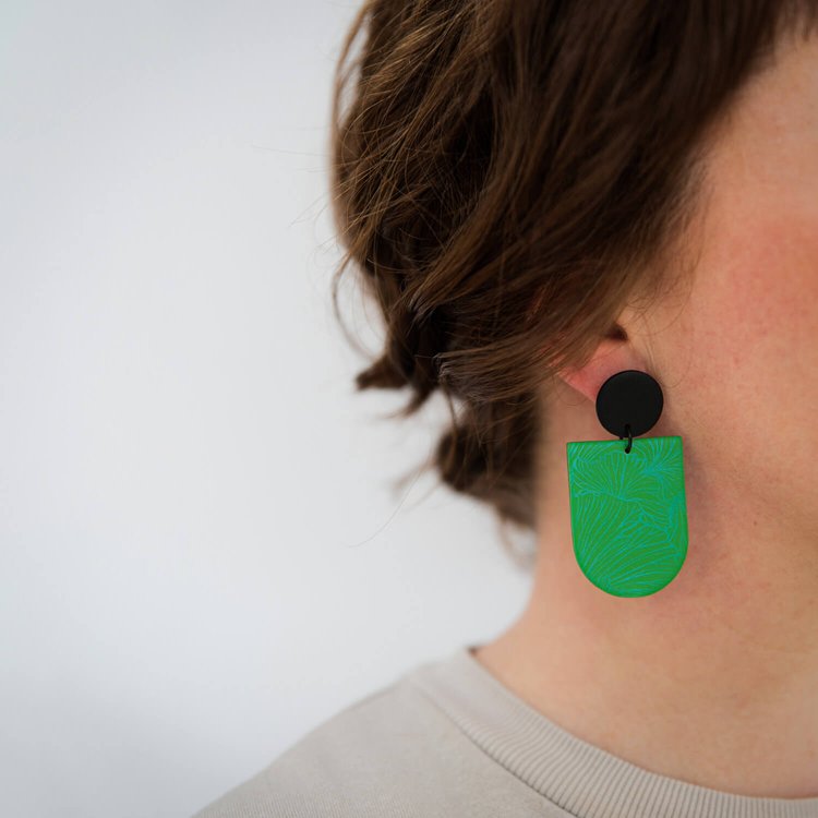Close-up of a person with wavy brown hair wearing the Cha Emerald Earrings by Culu Design—large geometric polymer clay earrings in emerald green and black. The background is plain white, and they are in a light beige top.