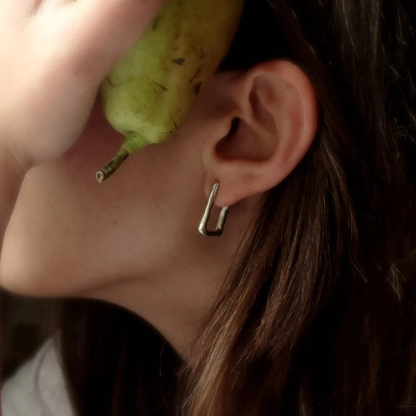 A person holds a pear near their face, drawing attention to their ear adorned with Inó Jewellery's Comfort Square Hoop Earrings. The sterling silver piece stands out against their brown hair, highlighting its sleek minimalist design.